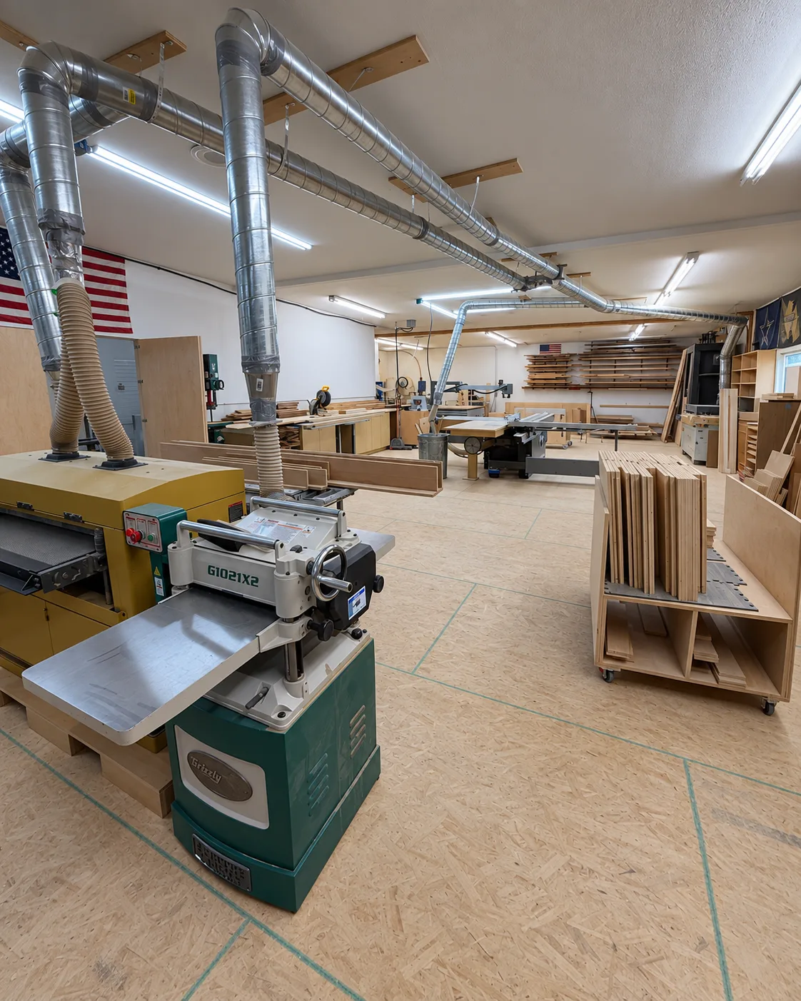 The shop floor in Ringgold: dust collection overhead, a thickness planer in front, a panel saw and stacks of cut parts behind.